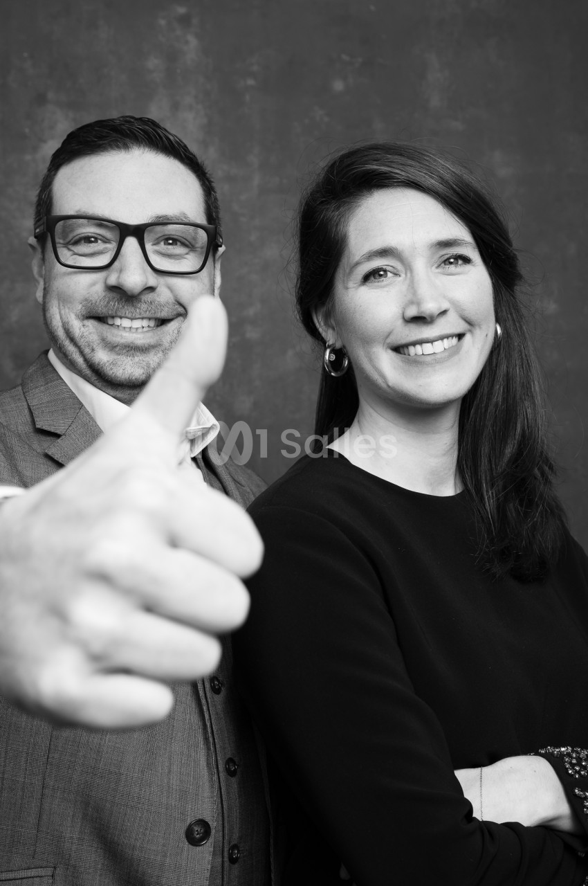 Portrait noir et blanc de deux personnes souriantes devant un fond sombre, l'homme au premier plan levant le pouce.