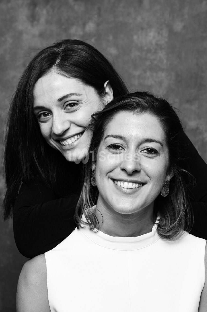 Portrait noir et blanc de deux femmes souriantes, l'une derrière l'autre, sur fond texturé gris.