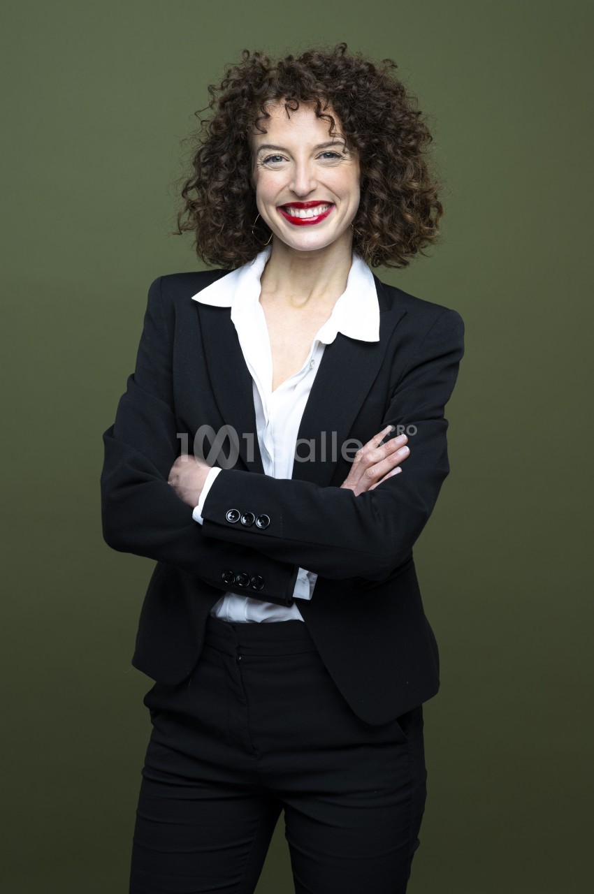 Portrait d'une femme aux cheveux bouclés, souriante, bras croisés, en tailleur noir sur chemise blanche, devant un fond vert uni.