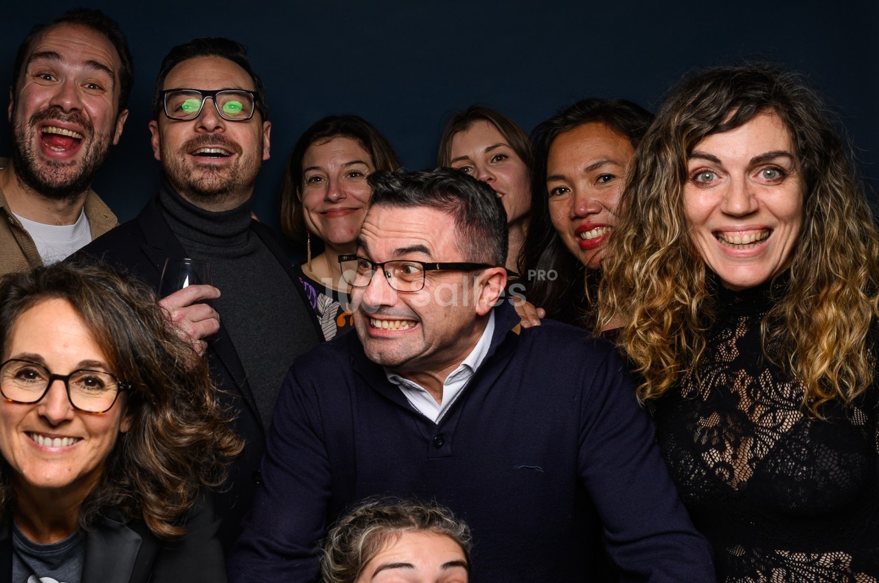 Groupe de huit personnes souriantes et posant serrées devant un fond bleu foncé, certaines tenant un verre, lors d'une séance photo fun. Let me check length: roughly 135 characters? Count: "Groupe de huit personnes souriantes et posant serrées devant un