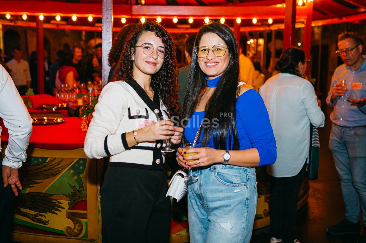 Deux femmes souriantes tenant un verre à cocktail, debout dans une salle animée aux guirlandes lumineuses rouges, avec des convives en arrière-plan.