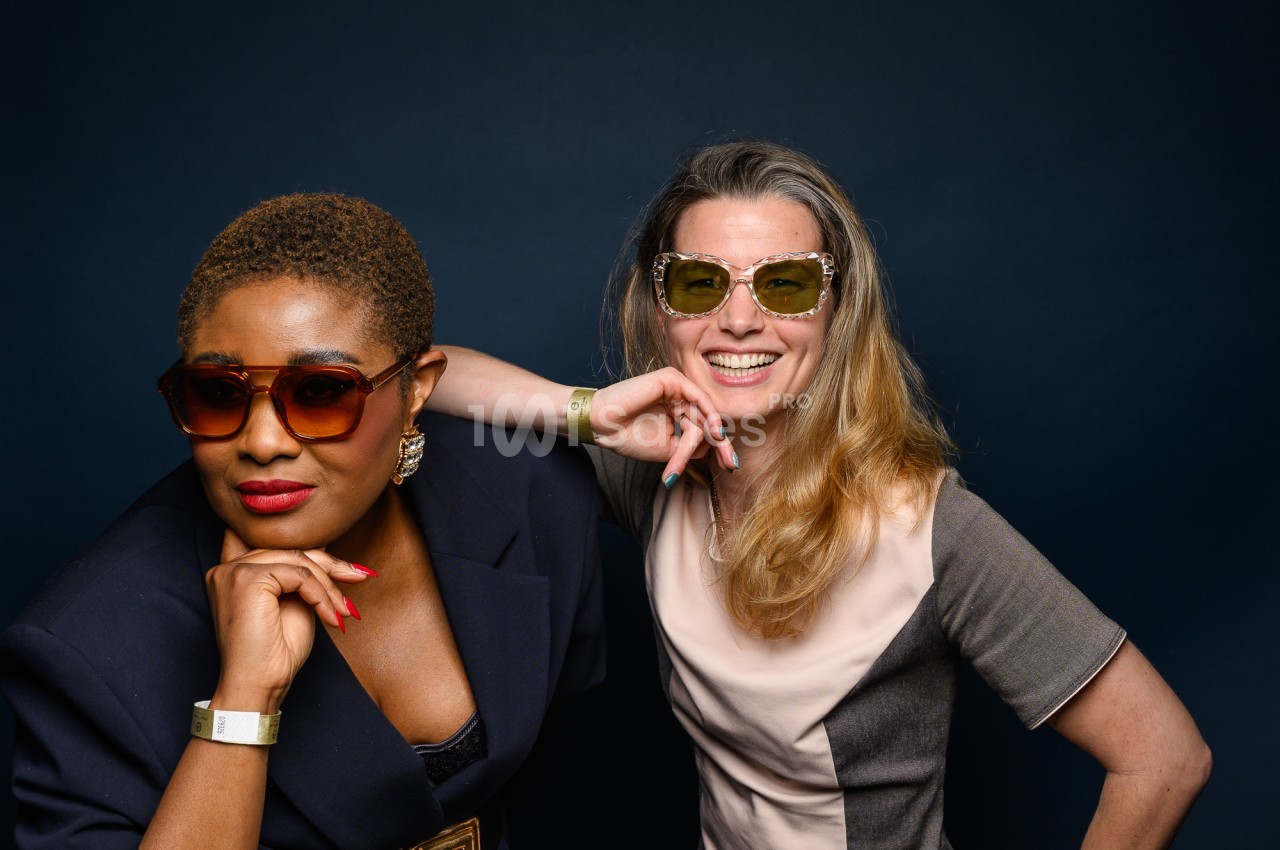 Deux femmes posent devant un fond bleu nuit, lunettes de soleil colorées, l'une en veste bleu marine et l'autre en robe bicolore, souriantes et complices.