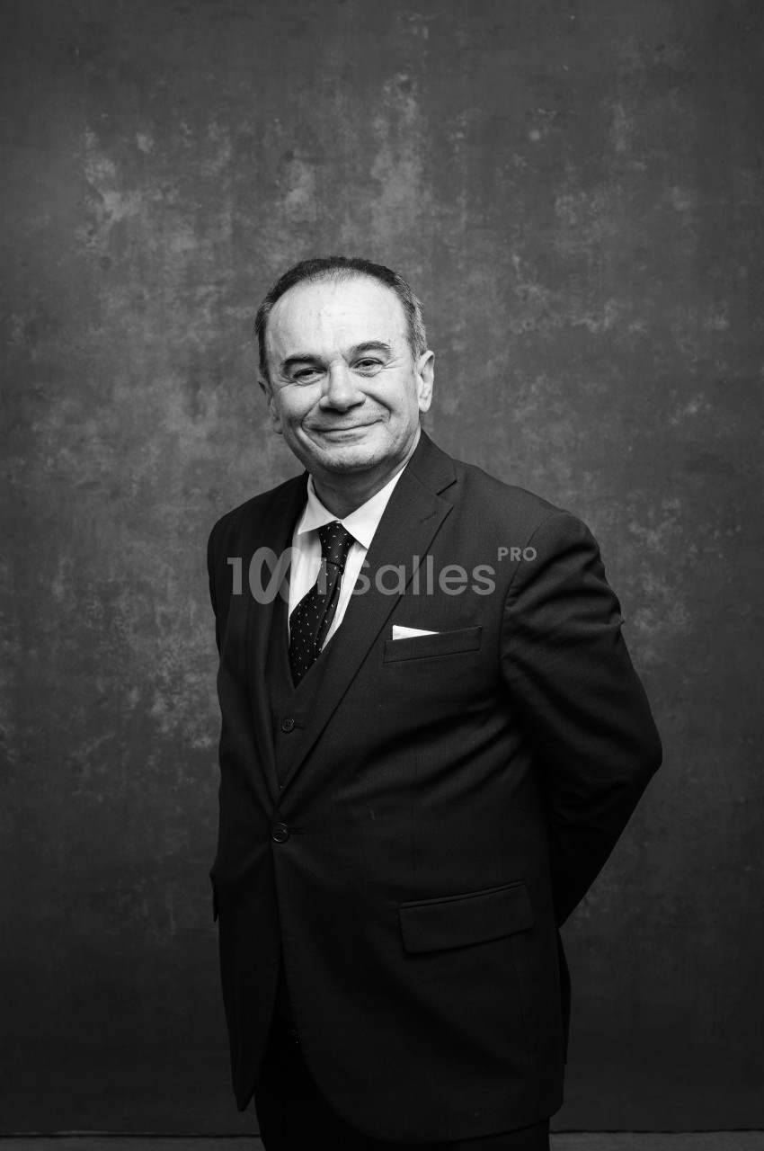 Portrait en noir et blanc d'un homme en costume sombre et cravate à pois, souriant, debout devant un fond gris texturé.