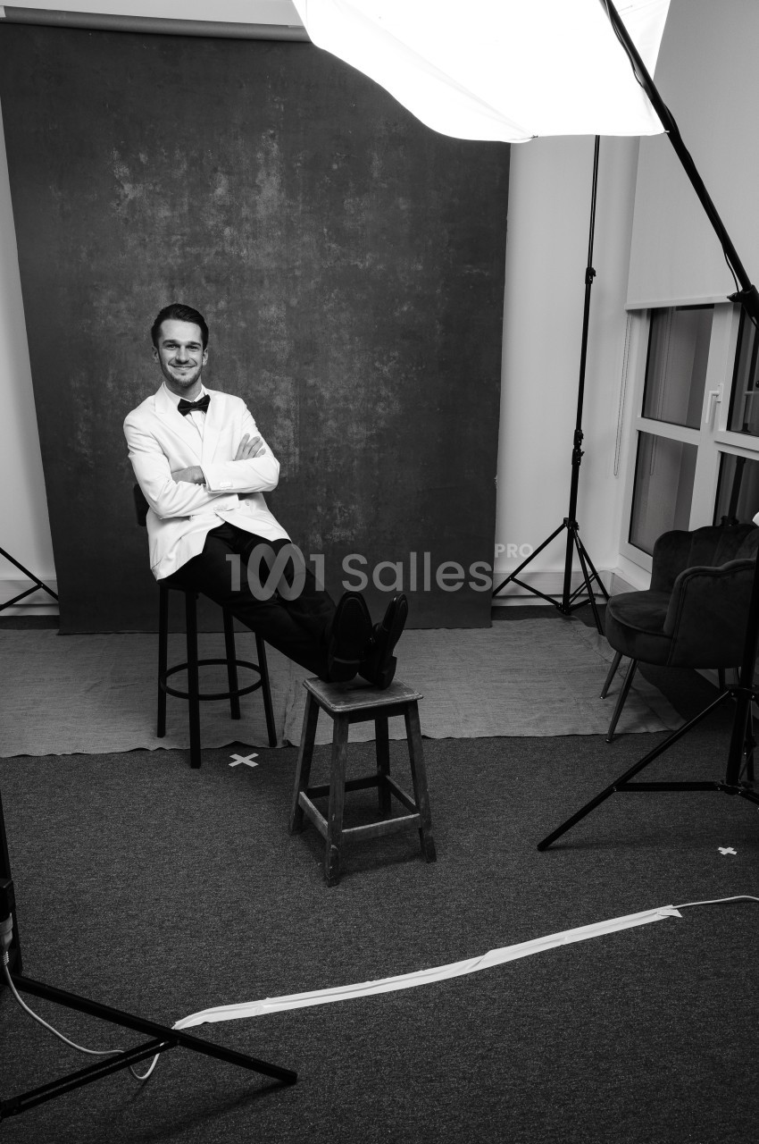 Homme souriant en veste blanche et nœud papillon, assis sur un tabouret de bar, jambes posées sur un petit tabouret en bois, dans un studio photo avec fond sombre texturé. Let me check length: roughly 165 characters, too long. Need ≤125. Homme souriant