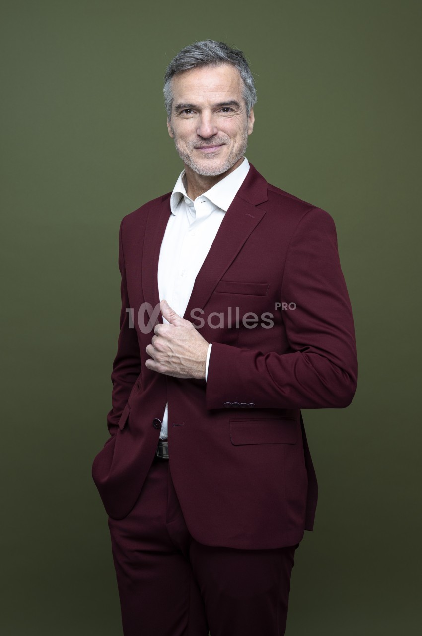 Homme aux cheveux gris en costume bordeaux et chemise blanche, debout devant un fond vert uni, une main posée sur le revers de sa veste.