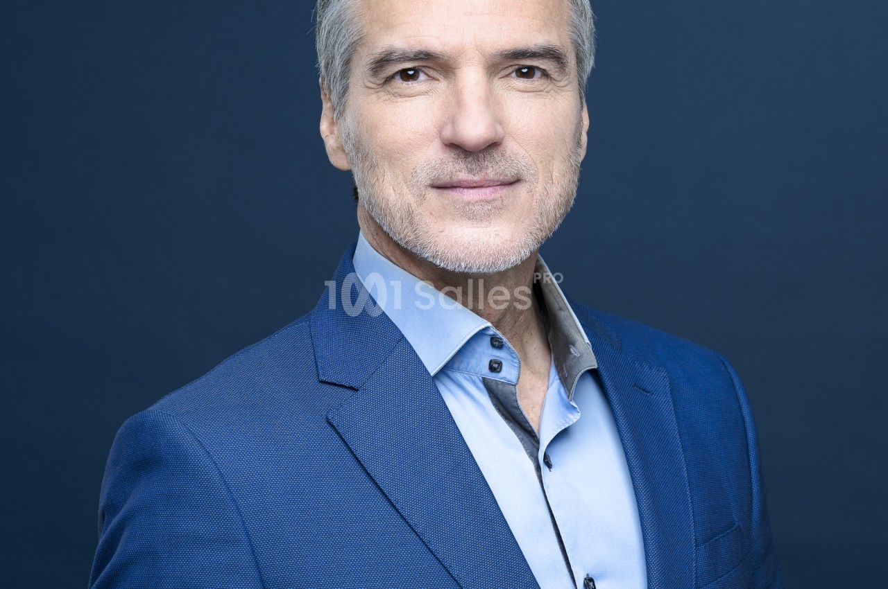 Portrait d'un homme aux cheveux gris, en veste bleue et chemise bleu clair, posant devant un fond bleu uni.