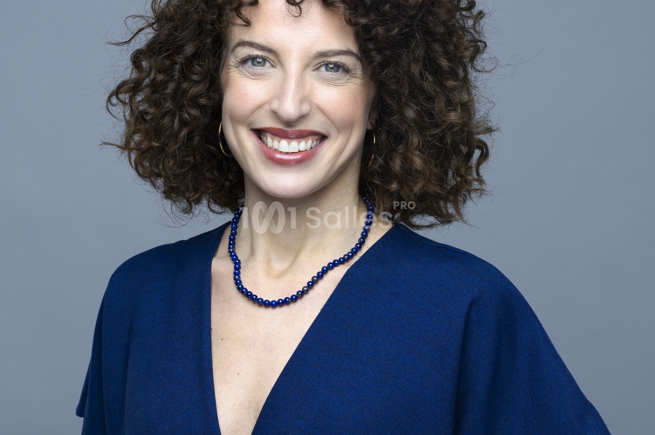 Portrait en gros plan d'une femme aux cheveux bouclés bruns, souriante, vêtue d'une robe bleu marine et d'un collier de perles bleues, sur fond uni gris.