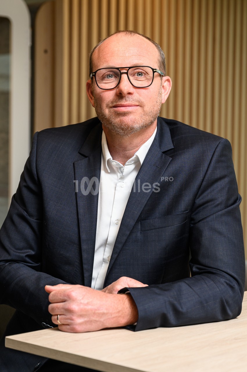 Homme d'âge mûr aux lunettes sombres, en veste bleu marine et chemise blanche, assis à une table claire devant un mur de lattes de bois.