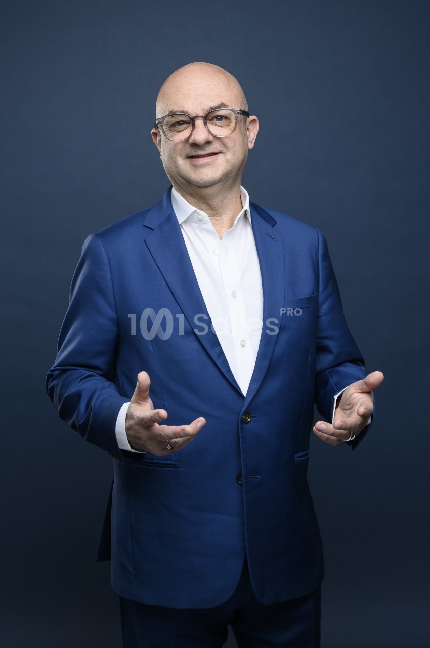 Homme chauve à lunettes au cadre clair, en costume bleu et chemise blanche, les mains ouvertes, posant devant un fond bleu uni.