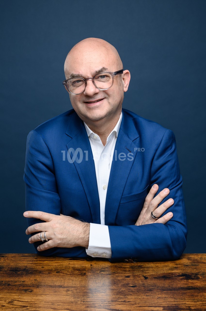 Homme chauve à lunettes à monture transparente, en veste bleu roi et chemise blanche, les bras croisés sur une table en bois, souriant face à l'objectif sur fond bleu uni.
