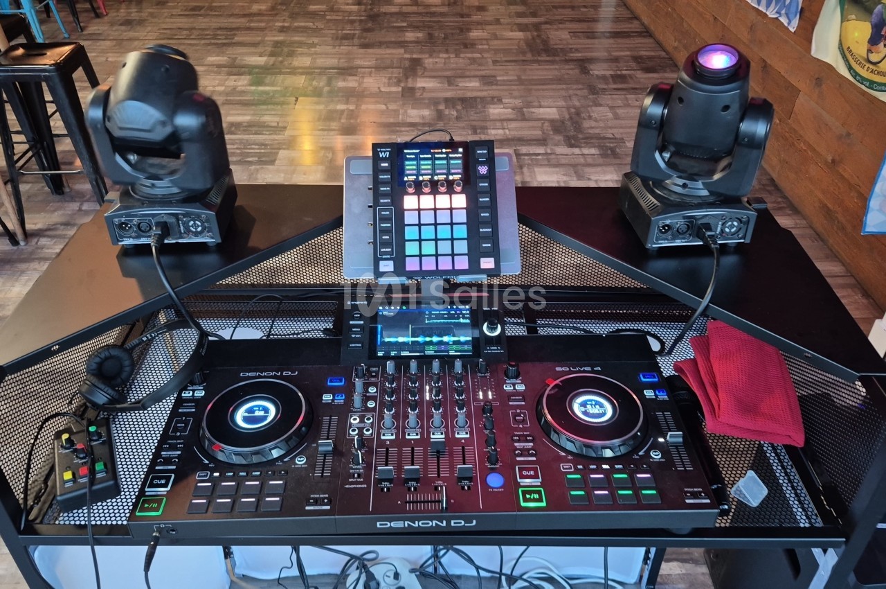 Console DJ Denon DJ SC Live 4 posée sur une table noire grillagée, avec deux lyres à tête mobile et un contrôleur de pads au centre, dans un lieu au sol en parquet.