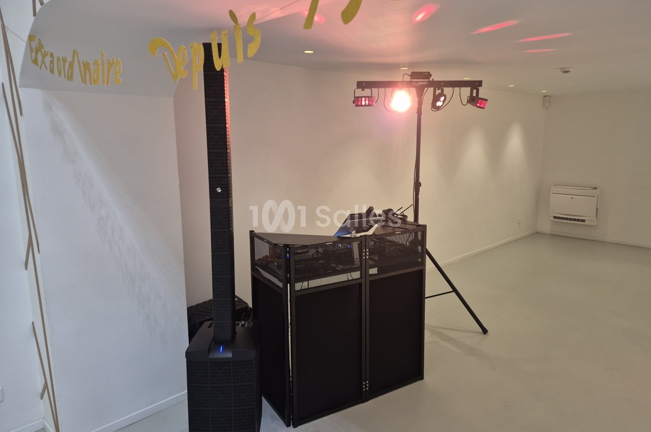 Pupitre de DJ noir avec grille métallique et colonne d'enceinte à côté, sous un jeu de projecteurs lumineux fixé sur une structure dans une pièce blanche vide. Let me check constraints: one sentence, under 125 chars? Count roughly: "Pupitre de DJ noir