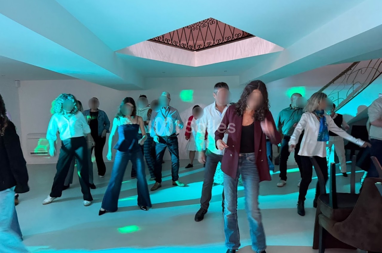 Des personnes dansent dans une salle aux murs blancs éclairée de lumières bleu-turquoise et vertes, sous un plafond en caisson avec une grille en fer forgé.
