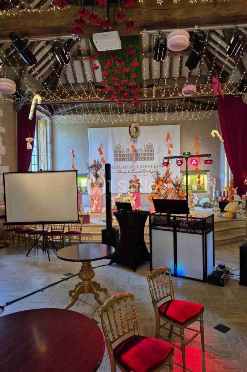 Salle aux poutres apparentes et guirlandes lumineuses, avec pupitre de DJ, écran de projection et chaises dorées.