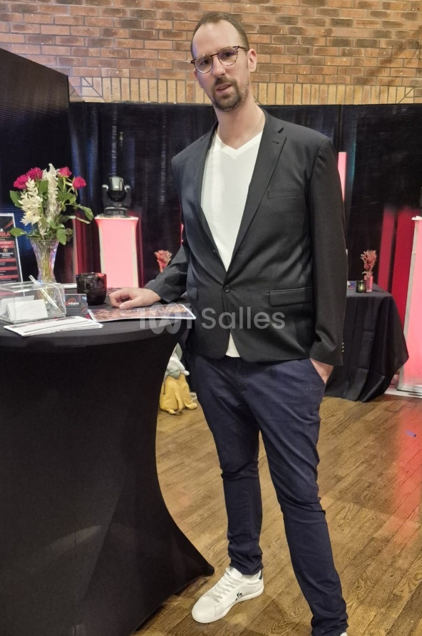 Homme en veste sombre debout près d'une table haute noire fleurie, dans une salle aux murs en briques.