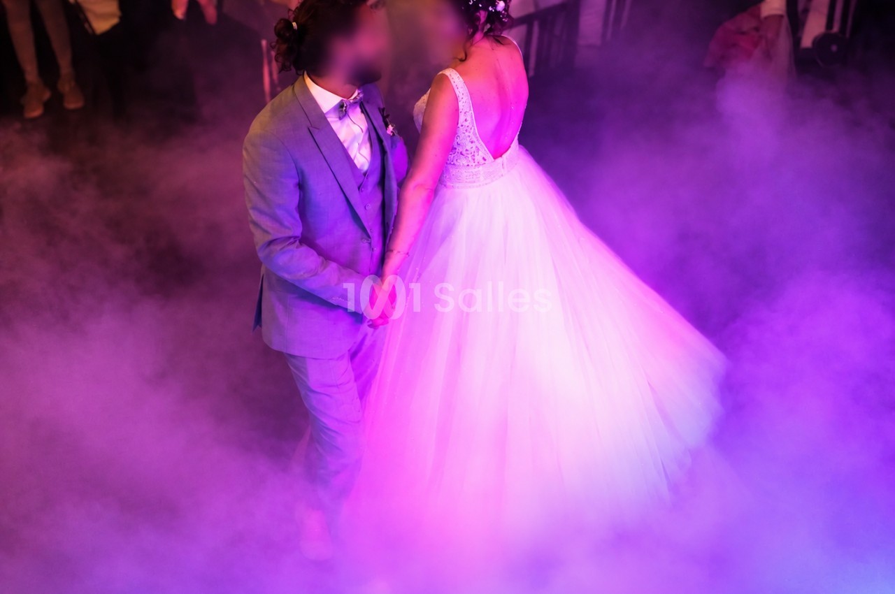 Des mariés dansent au centre d'une piste enveloppée de fumée, éclairée par des lumières violettes et roses ; la mariée porte une robe blanche à large jupe en tulle. Count characters: roughly 170 characters, too long. Need ≤125. Shorter: "Mariés dansant