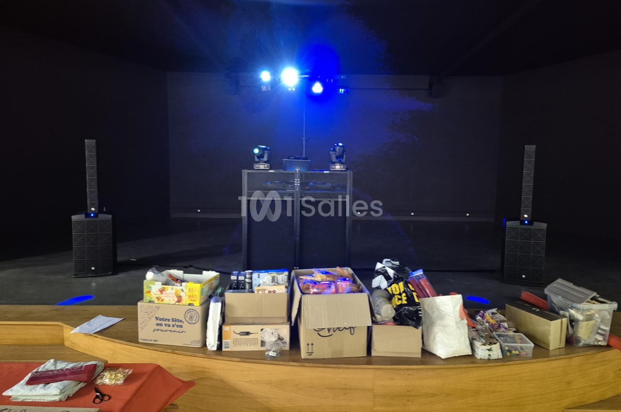 Scène sombre et plongée dans le bleu, avec deux projecteurs mobiles et deux enceintes colonnes de part et d'autre, une table de mixage au centre et des cartons de matériel posés sur le bord de scène.