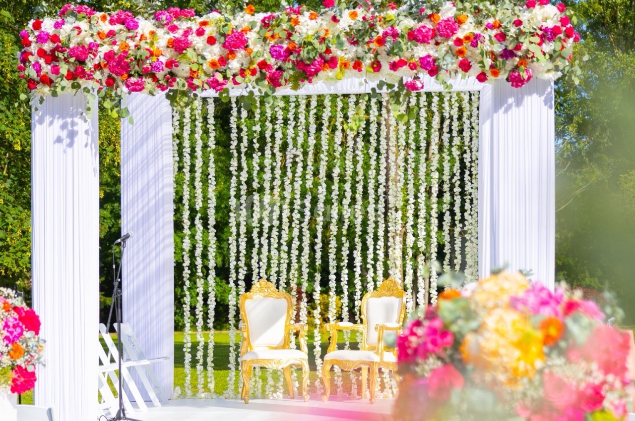 Estrade extérieure dressée de fleurs roses, rouges et orange, avec deux fauteuils dorés à dossier blanc sous une tenture de fleurs blanches retombantes.