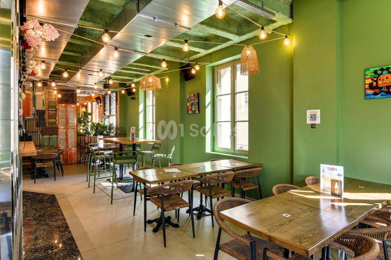 Salle de restaurant aux murs verts, avec tables en bois, chaises en osier, guirlandes d'ampoules suspendues et plafond végétalisé vert.