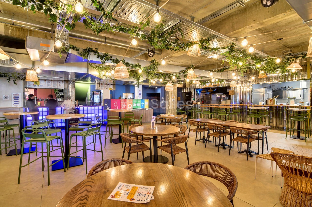 Espace de restauration aux tables en bois et chaises en rotin, sous une guirlande de guinguettes et des plantes suspendues, avec comptoirs et cuisine ouverte au fond. Let me check constraints: under 125 chars? Count roughly: "Espace de restauration aux