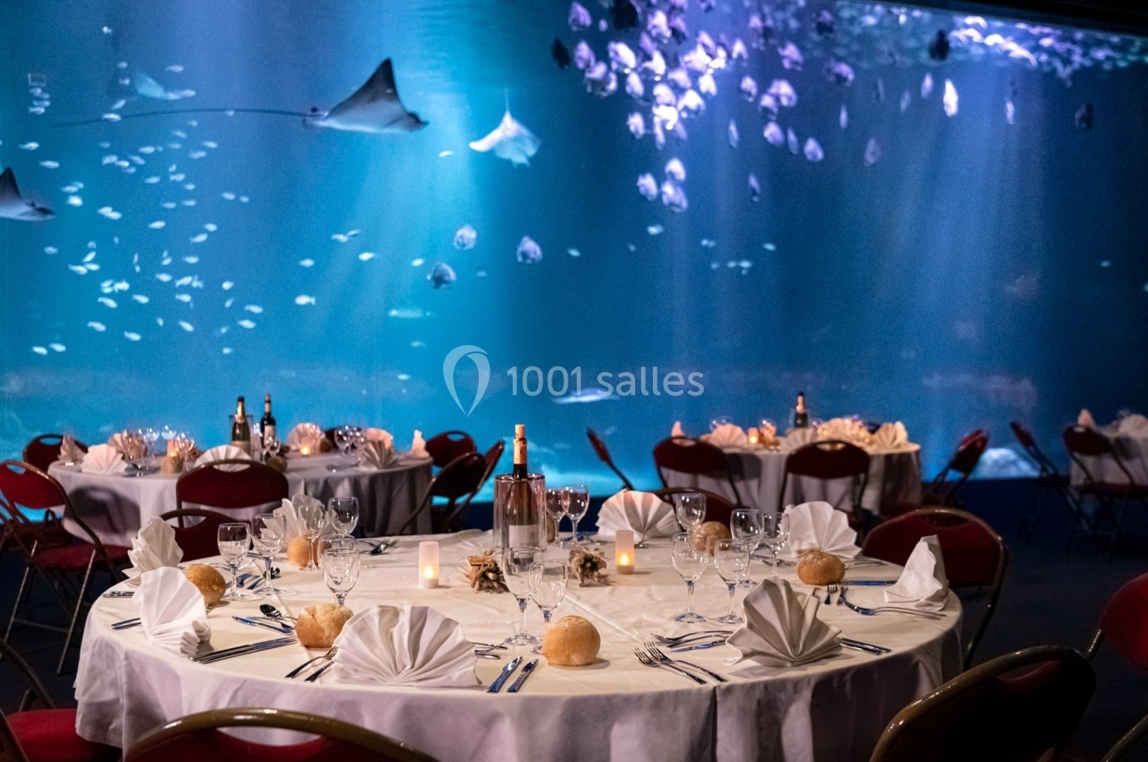 Salle de réception avec tables dressées, nappes blanches et vue sur un grand aquarium rempli de raies et poissons.