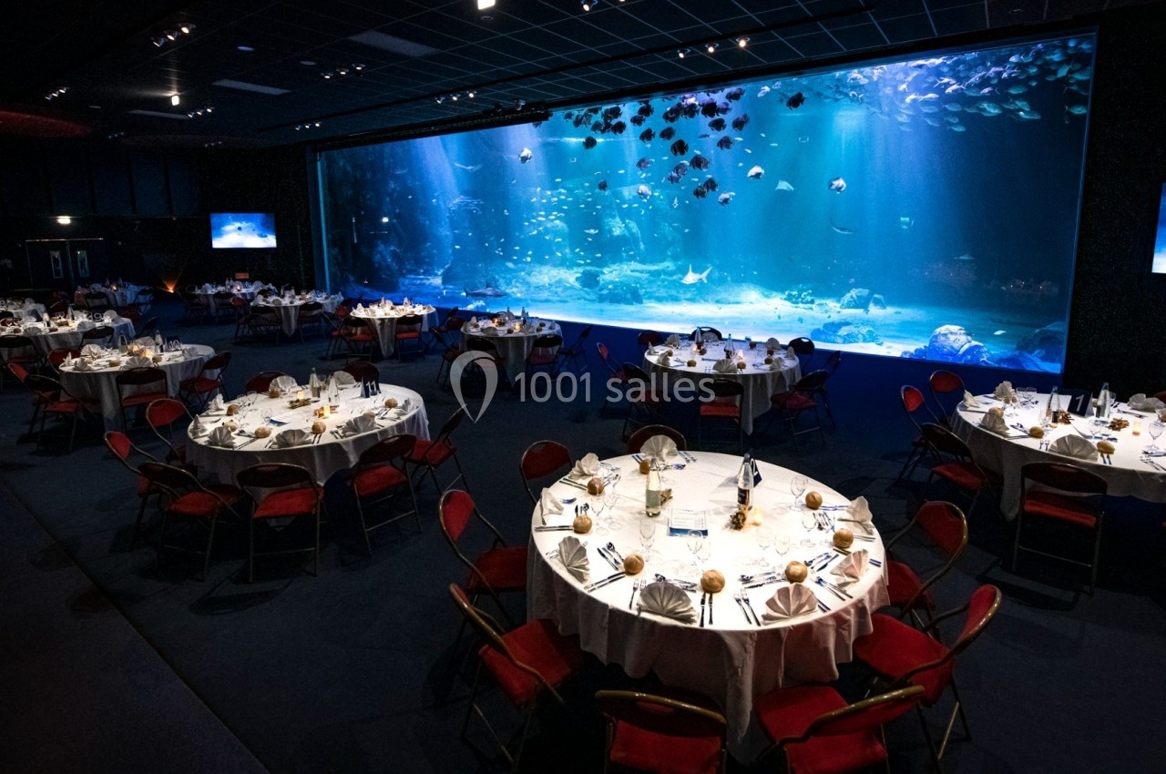Salle de réception avec tables rondes dressées, située devant un grand aquarium illuminé.