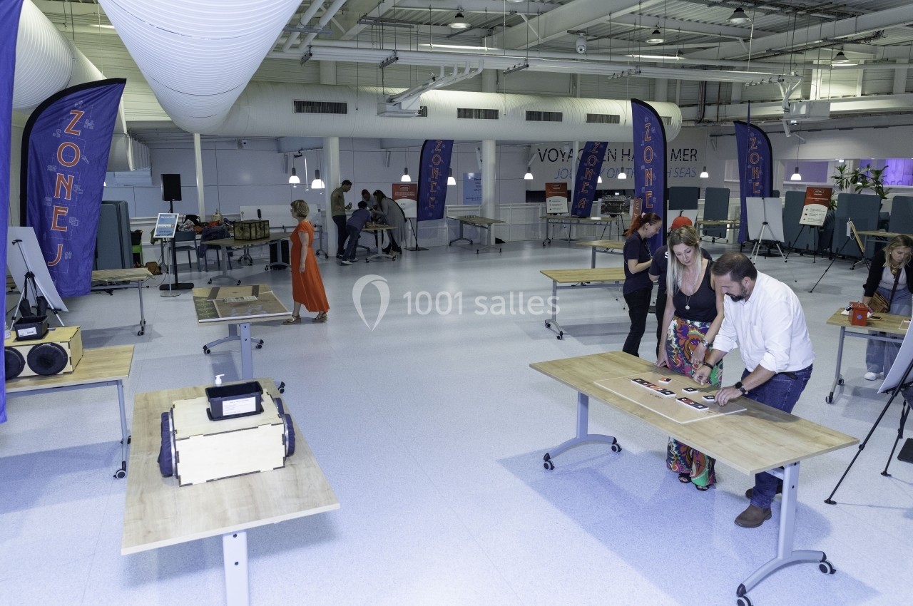 Salle lumineuse avec des tables en bois, des stands d'exposition et des personnes observant ou manipulant des objets.