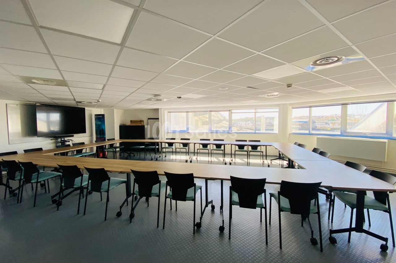 Salle de réunion lumineuse avec une grande table en U entourée de chaises noires, écran et fenêtres en arrière-plan.