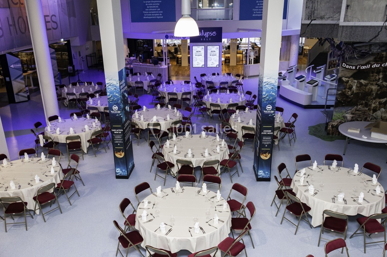 Salle aménagée avec des tables rondes dressées, entourées de chaises rouges, dans un espace lumineux et moderne.