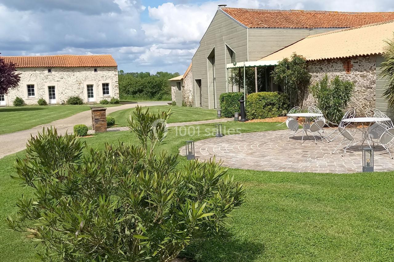 Cour extérieure avec tables et chaises sur une terrasse pavée, entourée de pelouse et bâtiments en pierre sous un ciel…