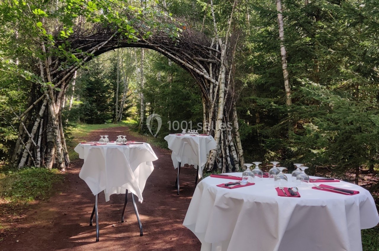 Tables dressées avec nappes blanches et couverts, disposées en plein air sous une arche en bois dans une forêt.