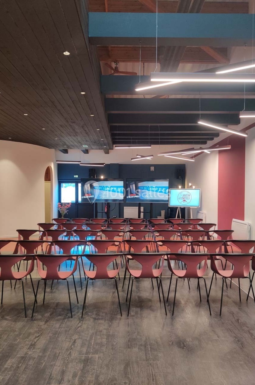Salle de conférence moderne avec des rangées de chaises rouges et des écrans allumés en fond.