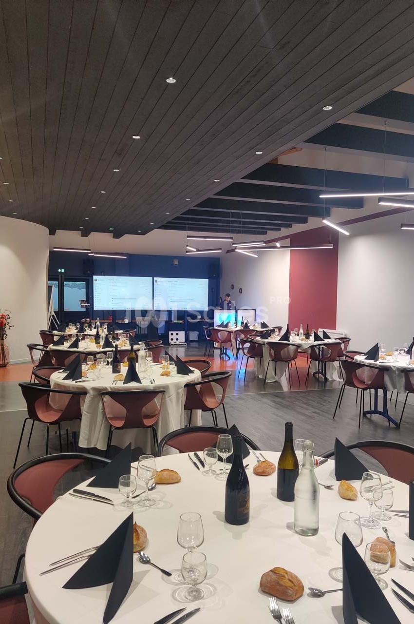 Salle de réception avec tables rondes dressées, nappes blanches, serviettes noires, bouteilles et écrans en arrière-plan.