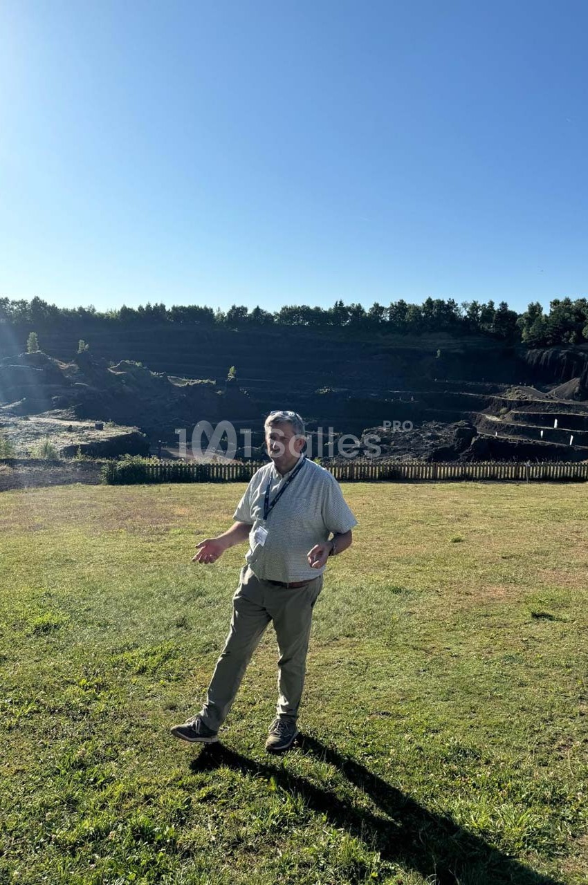 Un homme debout sur une pelouse ensoleillée, avec une carrière et des arbres en arrière-plan.