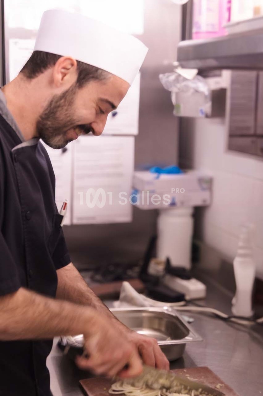 Un cuisinier en tenue professionnelle hache des ingrédients sur une planche dans une cuisine.