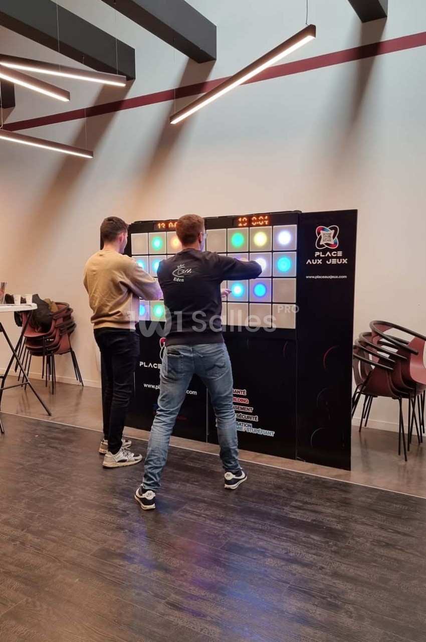 Deux personnes jouent sur un mur interactif lumineux dans une salle moderne avec des chaises empilées en arrière-plan.