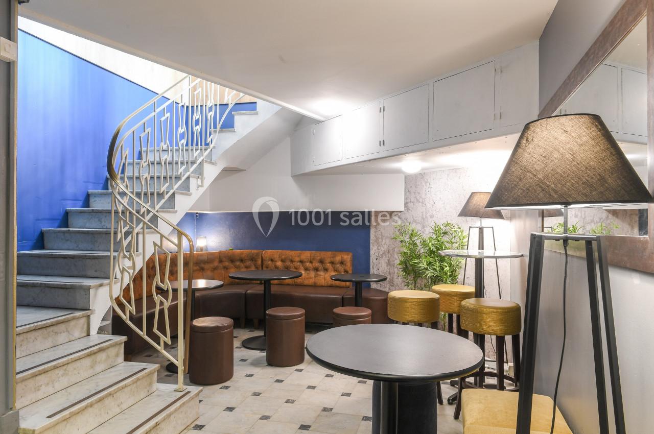 Espace intérieur avec escalier, tables rondes, tabourets, banquette en cuir et éclairage chaleureux.