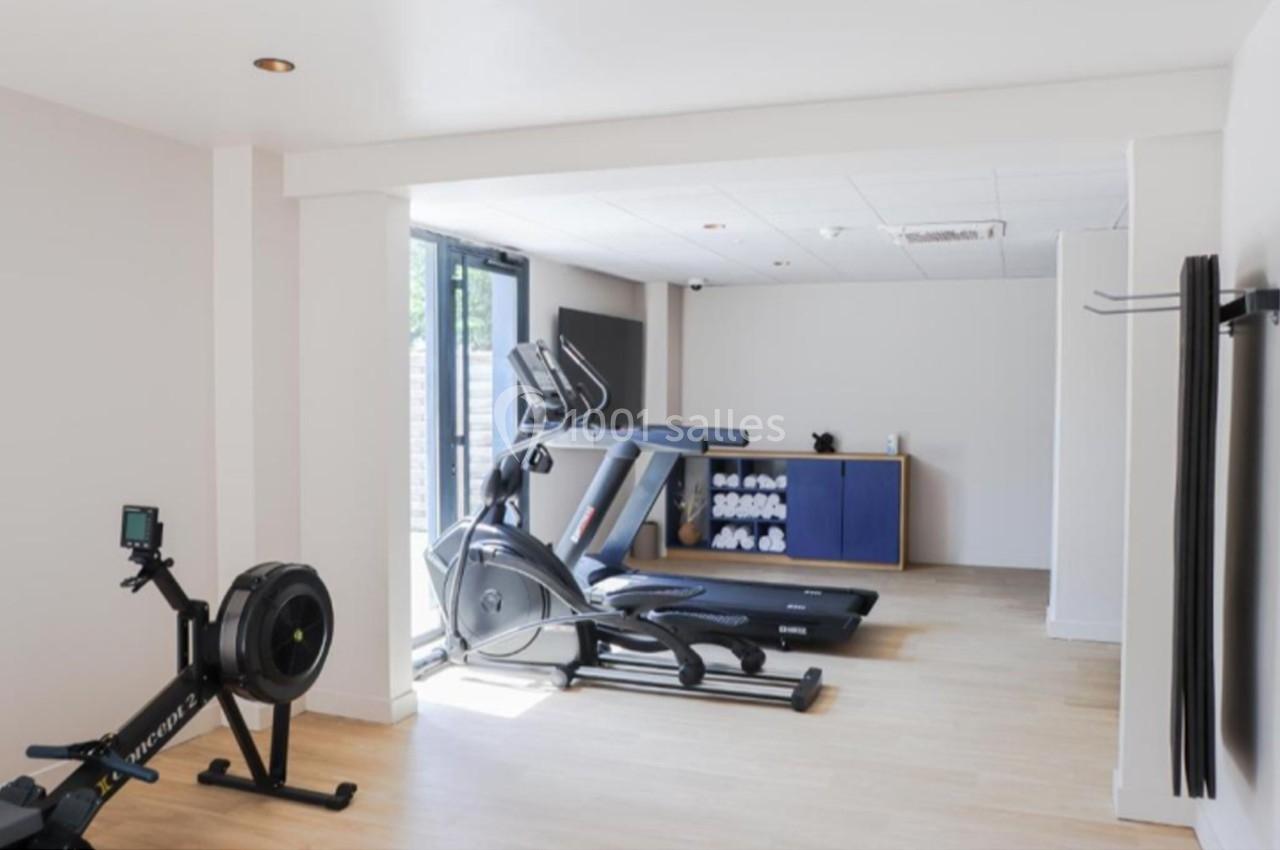 Salle de sport lumineuse avec tapis de course, rameur, étagère à serviettes et grande baie vitrée.