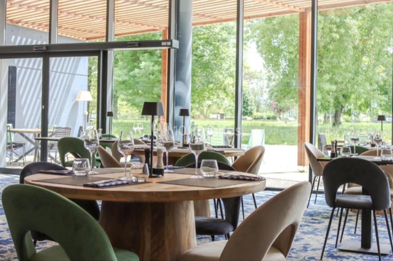 Salle de restaurant lumineuse avec grandes baies vitrées, tables en bois, chaises modernes et vue sur un jardin verdoyant.