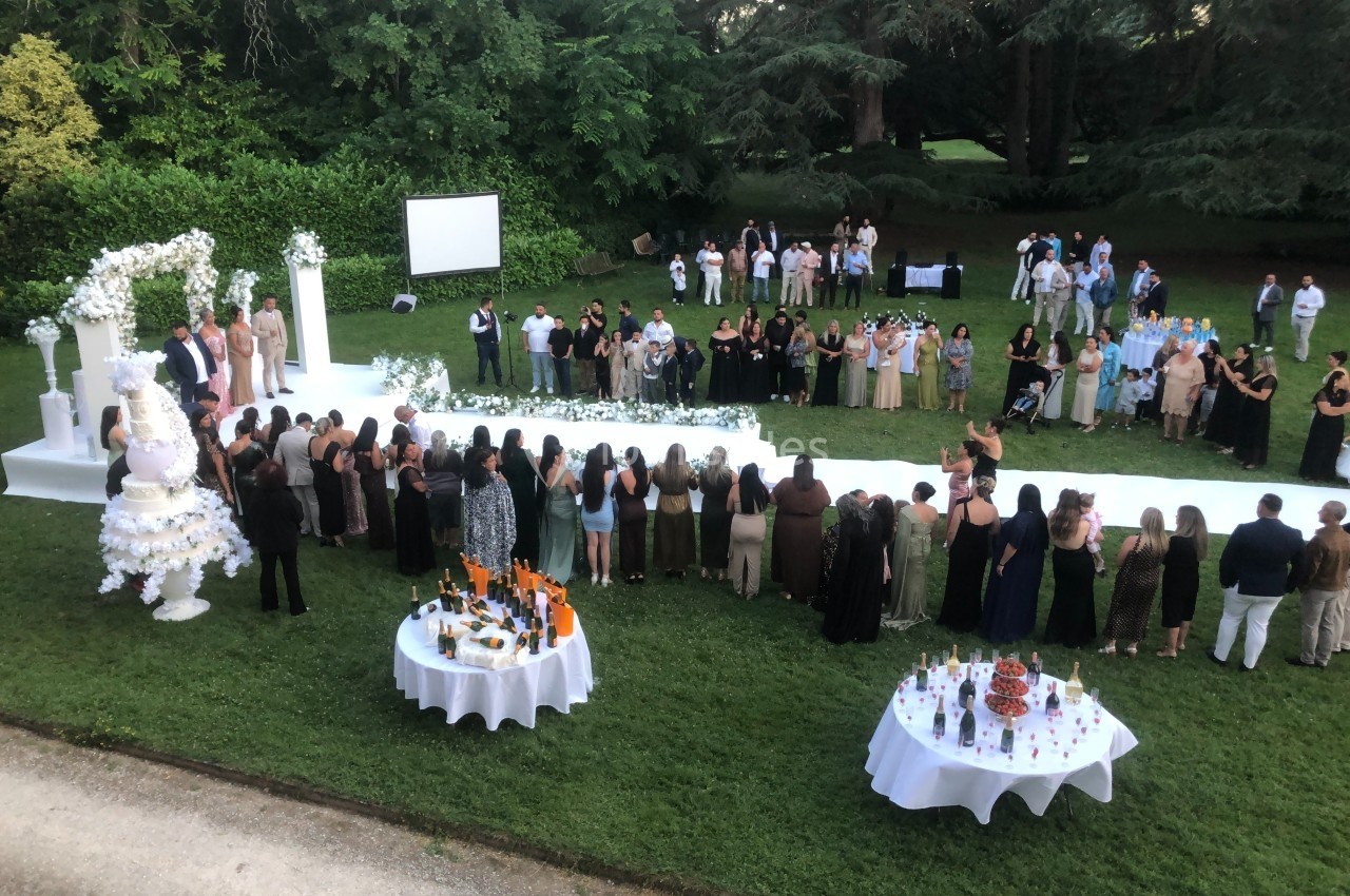 Groupe de personnes rassemblées dans un jardin pour un événement, avec tables dressées et décorations blanches.