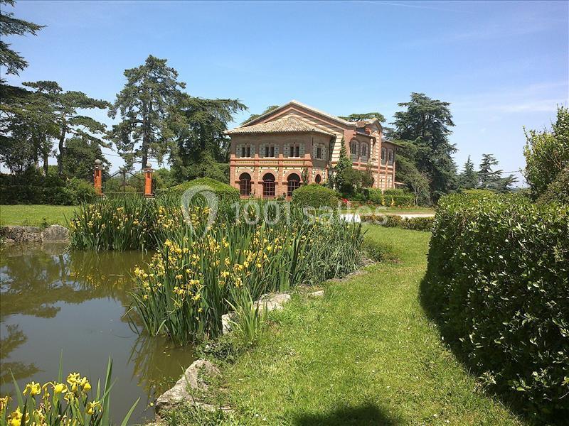 Manoir en briques rouges entouré de jardins verdoyants avec un étang bordé de roseaux au premier plan.