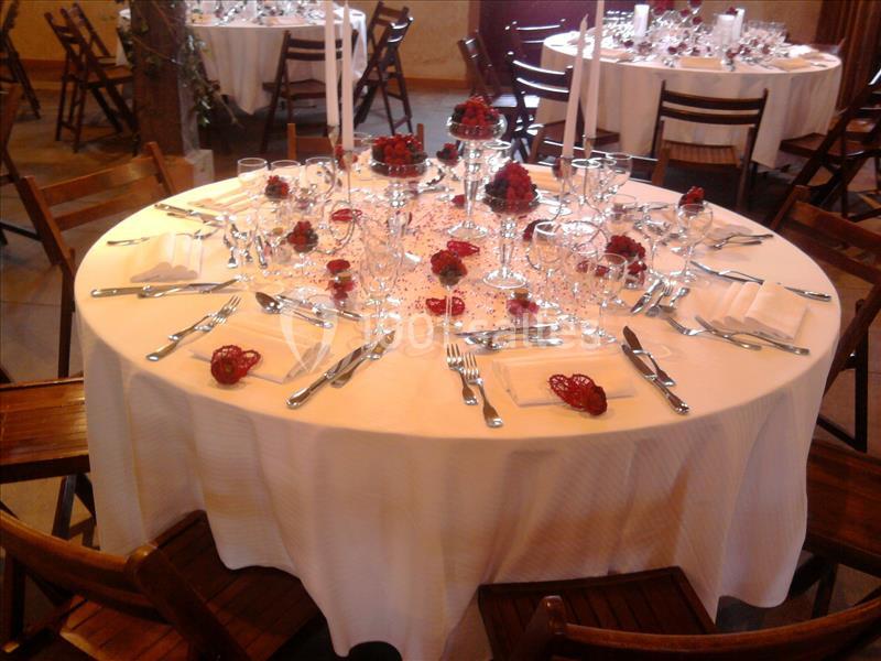 Table ronde élégamment dressée avec nappes blanches, couverts alignés, verres et décorations rouges.