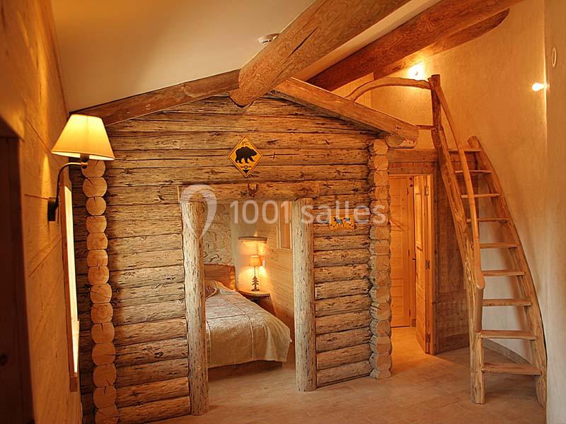 Chambre rustique avec murs en rondins, lit éclairé par une lampe, et mezzanine accessible par une échelle en bois.