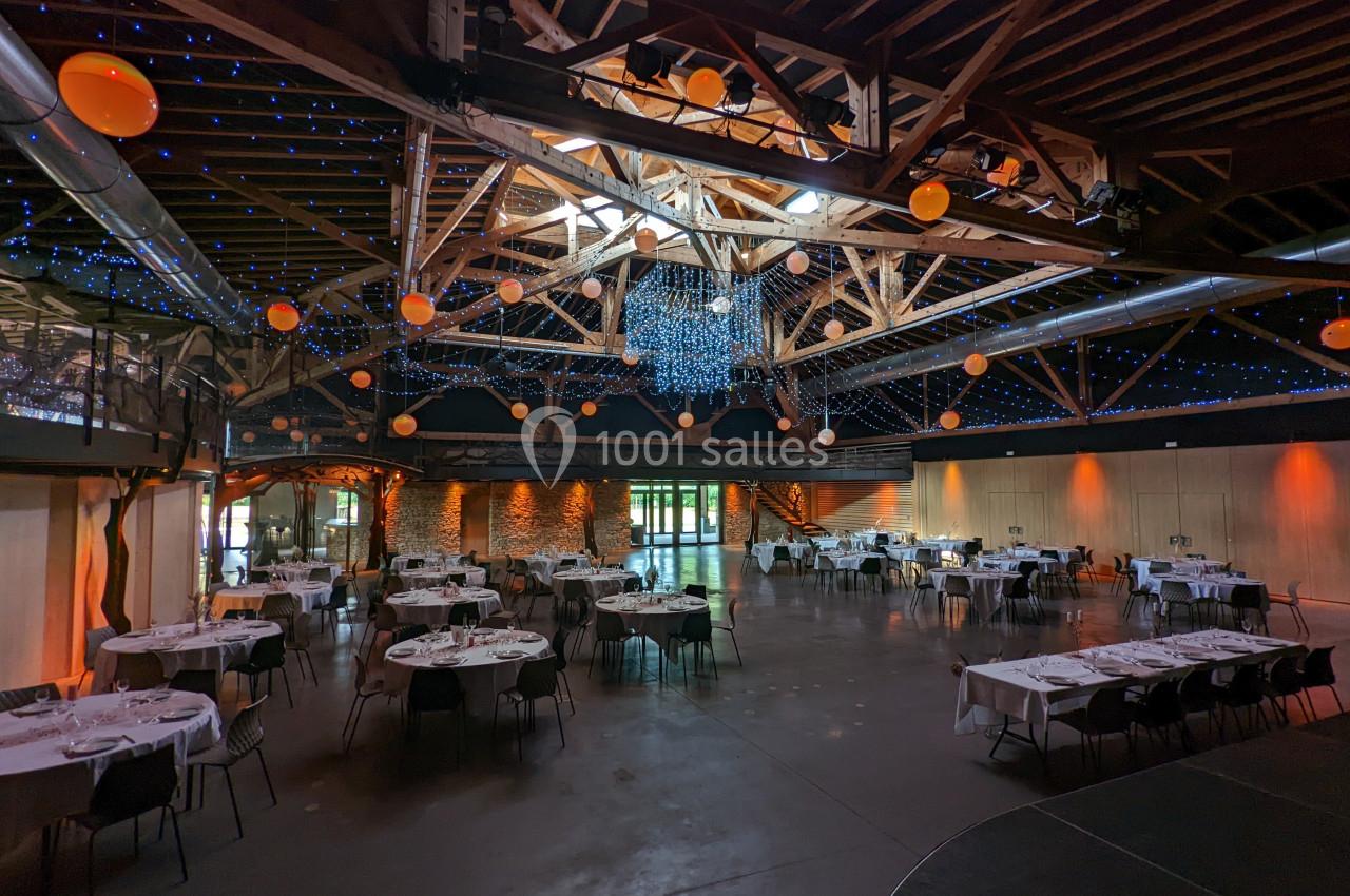 Salle de réception spacieuse avec tables dressées, poutres apparentes et éclairage décoratif suspendu.