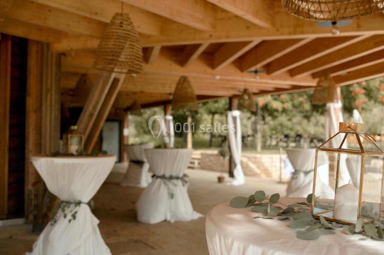 Salle de réception en extérieur avec tables hautes décorées de nappes blanches et lanternes suspendues en osier.