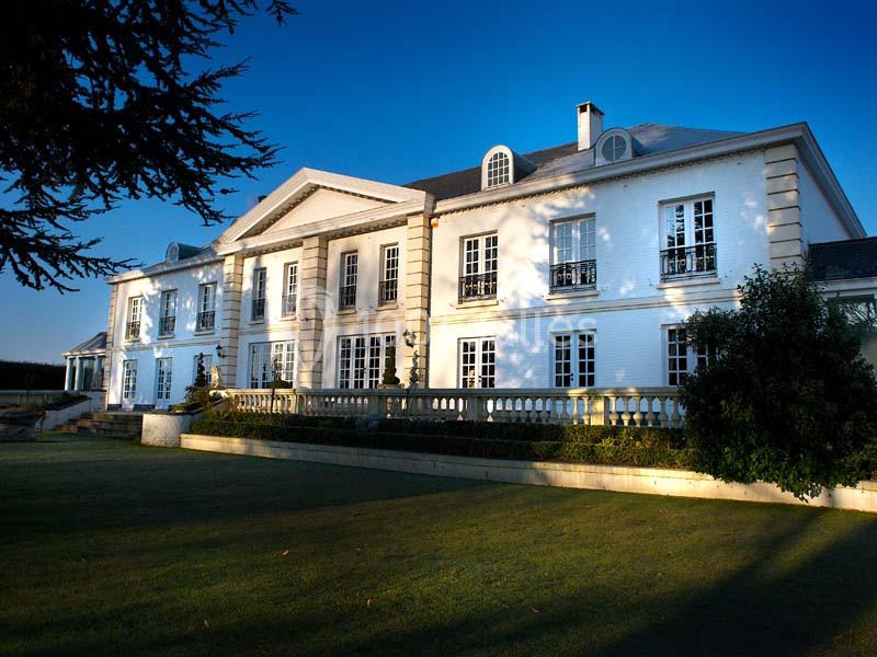 Façade d'un grand bâtiment blanc de style classique, entouré de pelouses et d'arbres sous un ciel bleu.