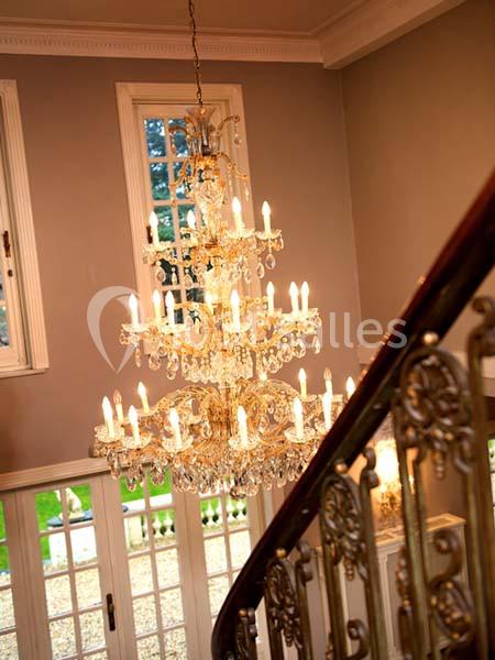 Lustre en cristal allumé suspendu dans une entrée avec escalier en fer forgé et grandes fenêtres.