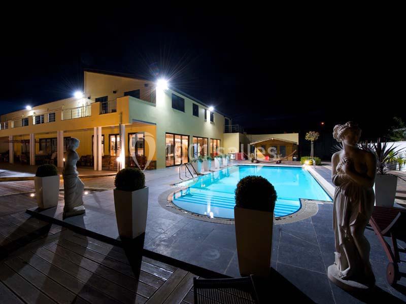 Vue nocturne d'une piscine éclairée entourée de statues et d'un bâtiment moderne avec de grandes baies vitrées.
