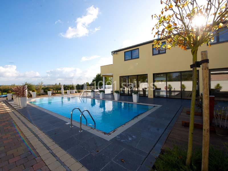 Piscine extérieure entourée de dalles grises, devant une maison moderne avec grandes baies vitrées.