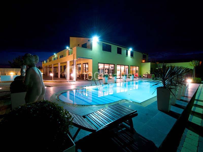 Vue nocturne d'une piscine éclairée devant un bâtiment moderne, entourée de plantes et de chaises longues.